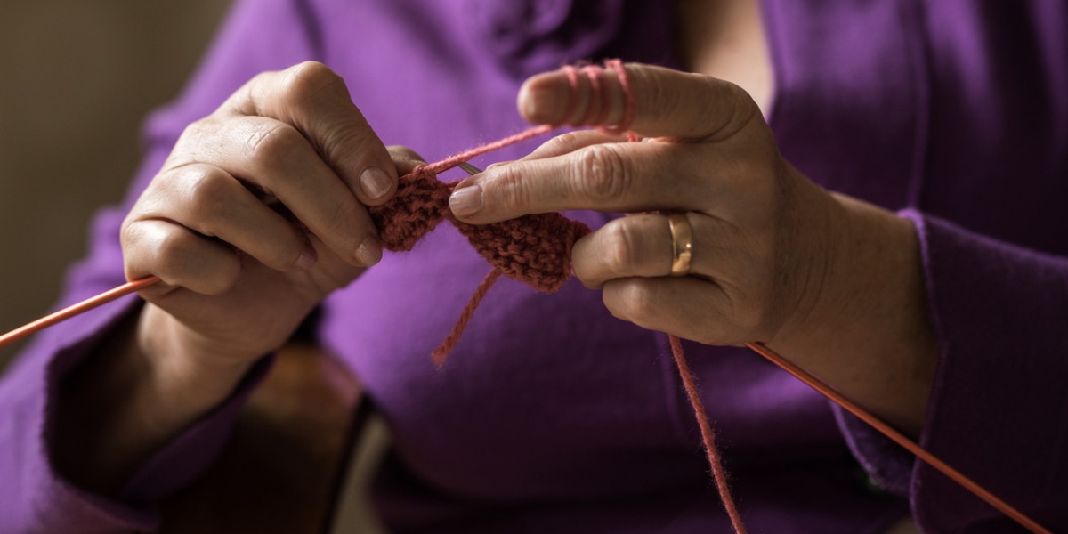 detail of older woman's hands knitting. she is wearing a purple jacket.