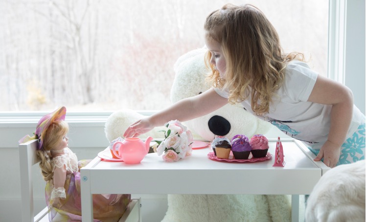 A little girl is having a tea party with her doll and teddy bear. On a white table there are flowers, a tea pot, plates and iced cupcakes.
