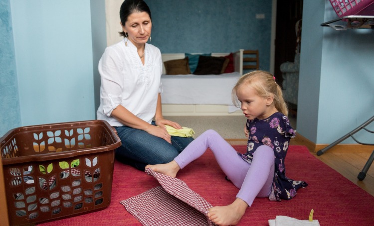 Girl with disability folds tea towel with her feet. Her mother watches on.