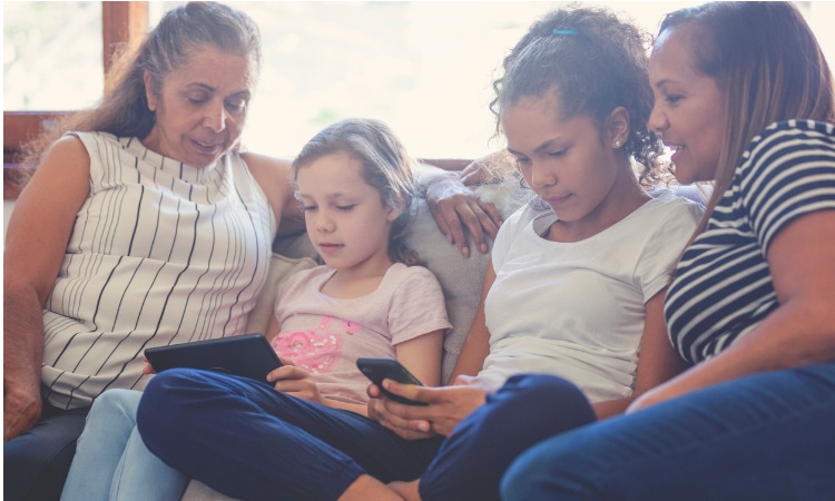 Image of multi-generational Indigenous family sitting on couch with the kids look at a smartphone and tablet device.