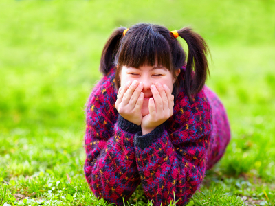 A young girl with disability is laying in the grass, she has her eyes closed, is smiling with her hands partly covering her face.