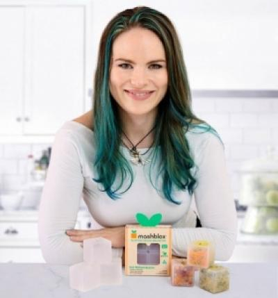 A woman with dark and green hair, smiling and sitting behind a Mashblox product display