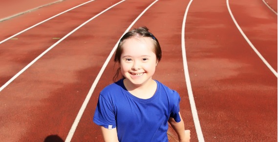 A photo of a young girl with down syndrome smiling at the camera. She is standing on an athletics track.