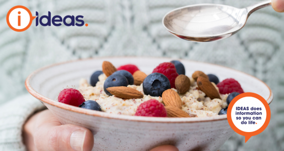An image of a person in a light grey winter jumper. In one hand they hold a speckled bowl, inside the bowl is porridge, topped with raspberries, blueberries and almonds. In the other hand they hold a silver spoon over the bowl.