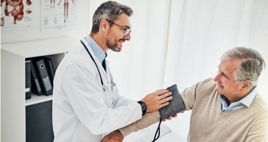 A Doctor wearing white coat and glasses has a stethoscope around his neck. He is holding a blood pressure monitor and wrapping the band around a patients arm in preparation to take blood pressure reading.