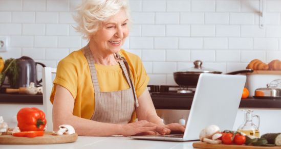 An image of an older woman in a kitchen looking at a laptop. She is wearing an apron and is surrounded by vegetables and cooking utensils
