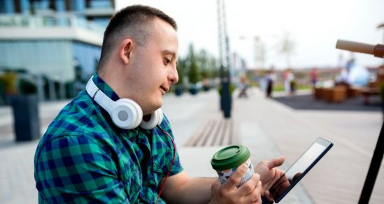 A teen male living with intellectual disabilty, sitting looking at a tablet device, He is holding a reusable takeaway cup and has headphones resting on his neck.of
