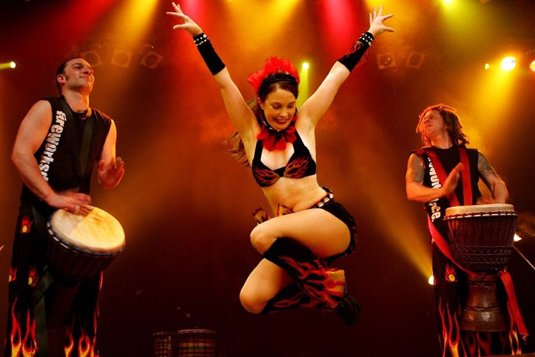 Woman dancing on stage with two drummers. She is wearing a bikini top and black boots with red and yellow flame detailing.
