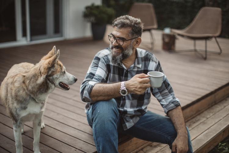 Image of a sitting man, holding a cup and looking at a dog.
