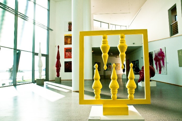 Art gallery featuring a yellow art exhibit displayed in the middle of the floor. Other exhibits are hung on the walls and surrounding areas